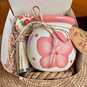 May include: A gift box containing a hand-painted ceramic candle holder with a pink floral design, a small glass bottle of matches, and a round cookie. The candle holder is tied with twine and has a tag that reads "Scented Candle".