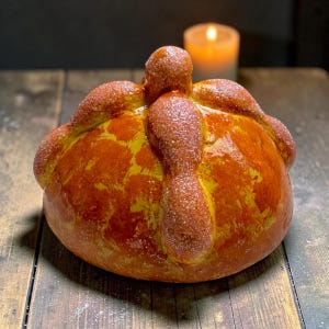 May include: A round, golden-brown pan de muerto bread featuring a central knob and four radiating bone-shaped dough strips on top. The surface is glazed and coated with a light dusting of granulated sugar, creating a textured, sweet appearance.