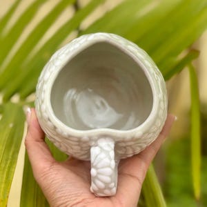 Puede incluir: Una taza de cer&aacute;mica verde claro hecha a mano con un patr&oacute;n texturizado y punteado. La taza tiene una abertura en forma de coraz&oacute;n y un asa a juego. La taza se sostiene en la mano de una persona, con un fondo verde borroso.