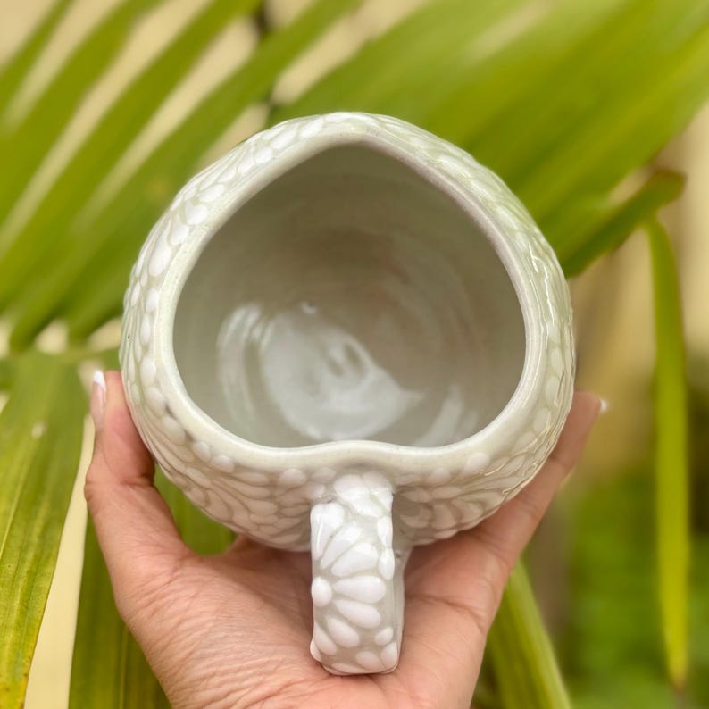 Puede incluir: Una taza de cer&aacute;mica verde claro hecha a mano con un patr&oacute;n texturizado y punteado. La taza tiene una abertura en forma de coraz&oacute;n y un asa a juego. La taza se sostiene en la mano de una persona, con un fondo verde borroso.