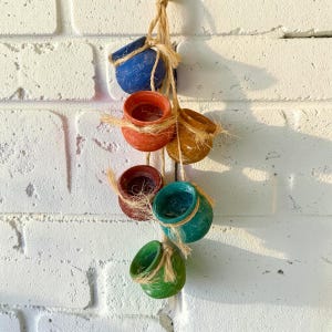 May include: A hanging decoration featuring six small, round, ceramic pots in blue, orange, yellow, dark red, teal, and green. Each pot is suspended by natural-colored twine, creating a vertical cluster of colorful, open-topped vessels.
