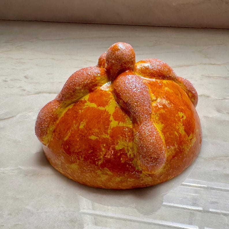 May include: A round, golden-orange pan de muerto bread featuring a central knob and four bone-shaped dough strips radiating from the top. The surface has a glossy, textured finish with a light dusting of sparkling sugar crystals throughout.