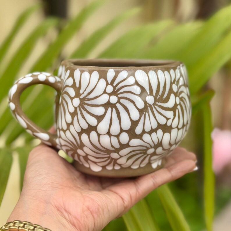Puede incluir: Taza de cer&aacute;mica marr&oacute;n con un dise&ntilde;o floral blanco. La taza, sostenida en una mano, tiene forma redondeada y un asa. El dise&ntilde;o floral blanco cubre el exterior de la taza. Adecuada para bebidas calientes, es decorativa.