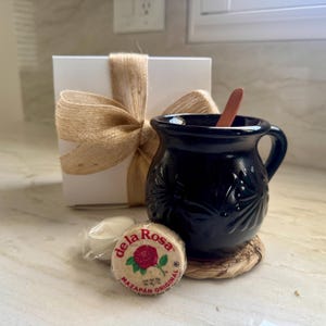 May include: A black ceramic mug with floral detailing, a wooden spoon, and a round mazapan candy with the text "de la Rosa". A white gift box tied with a burlap ribbon is in the background. A small, wrapped item is also present.