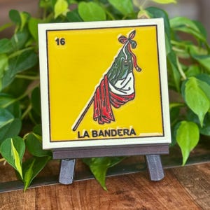 May include: A decorative tile featuring a vibrant yellow background with a flag design. The tile has a white border and the number 16 in the upper left corner. The text "LA BANDERA" is at the bottom. The tile is displayed on a small wooden easel.