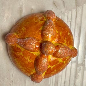 May include: A round, golden-orange bread-shaped object featuring a central knob and four radiating bone-like protrusions. The surface has a glossy, textured finish with a light dusting of fine, sparkling glitter across the entire top structure.