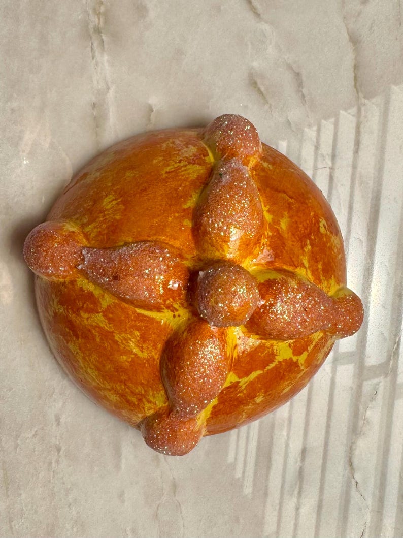 May include: A round, golden-orange bread-shaped object featuring a central knob and four radiating bone-like protrusions. The surface has a glossy, textured finish with a light dusting of fine, sparkling glitter across the entire top structure.