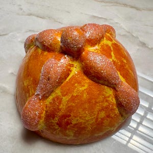 May include: A round, golden-orange bread roll featuring a central knob and four radiating bone-shaped dough pieces on top. The surface has a glossy, baked finish with a light dusting of sparkling, clear glitter particles across the entire structure.