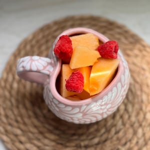 Puede incluir: Una taza rosa en forma de coraz&oacute;n llena de trozos de mel&oacute;n y frambuesas frescas. La taza tiene un dise&ntilde;o floral blanco y un asa a juego. La fruta est&aacute; dispuesta en la taza sobre una estera tejida.