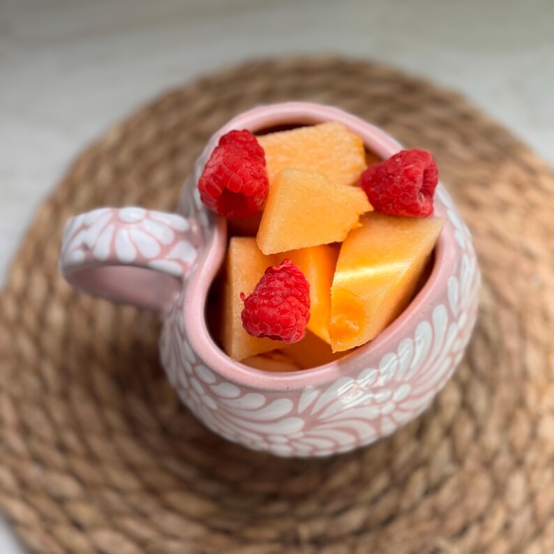 Puede incluir: Una taza rosa en forma de coraz&oacute;n llena de trozos de mel&oacute;n y frambuesas frescas. La taza tiene un dise&ntilde;o floral blanco y un asa a juego. La fruta est&aacute; dispuesta en la taza sobre una estera tejida.
