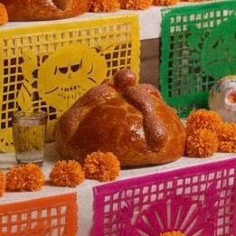 May include: A round, golden-brown pan de muerto bread with a central knob and bone-shaped dough decorations, placed on a tiered surface with orange marigold flowers, a small glass of clear liquid, and yellow, green, and pink papel picado banners.