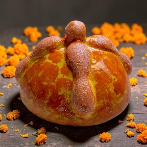May include: A round, golden-brown pan de muerto bread featuring a central knob and decorative bone-shaped dough strips on top, all lightly dusted with sparkling sugar crystals and surrounded by scattered bright orange marigold flower petals.