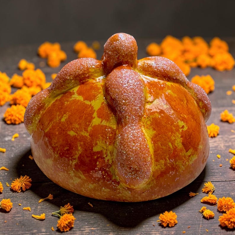 May include: A round, golden-brown pan de muerto bread featuring a central knob and decorative bone-shaped dough strips on top, all lightly dusted with sparkling sugar crystals and surrounded by scattered bright orange marigold flower petals.