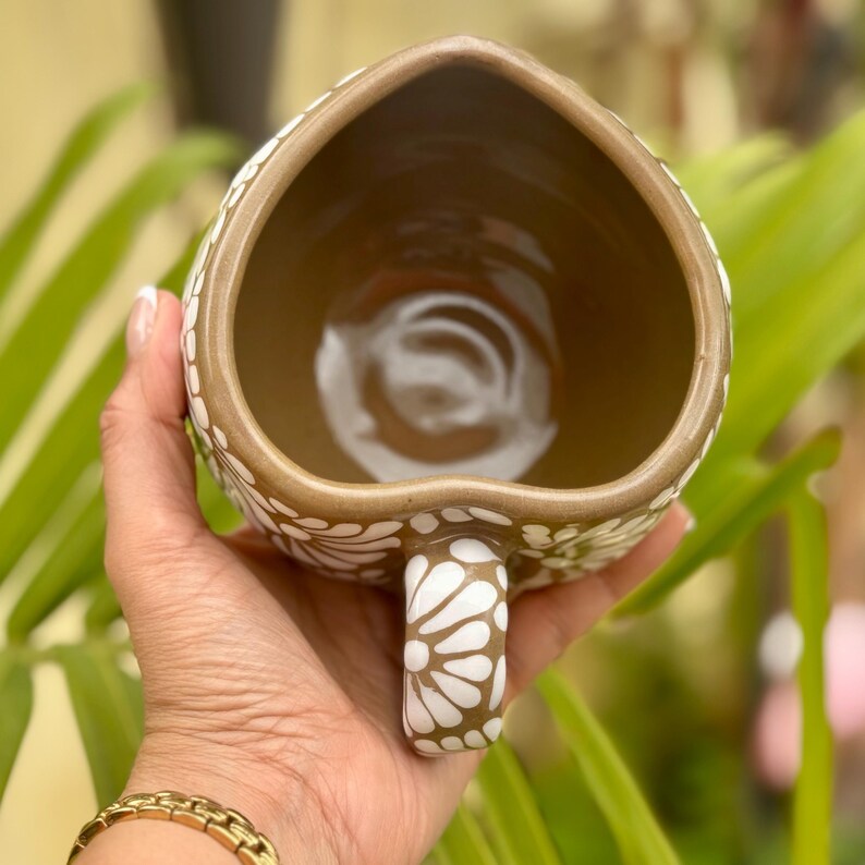 Puede incluir: Una taza de cer&aacute;mica en forma de coraz&oacute;n con interior y exterior marr&oacute;n. El exterior est&aacute; decorado con un estampado floral blanco. El asa tambi&eacute;n est&aacute; decorada con el mismo estampado floral blanco. La taza se sostiene en una mano.
