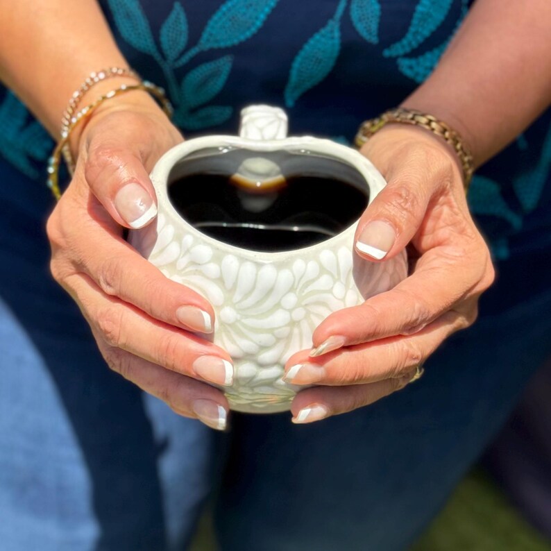 Puede incluir: Una taza de cer&aacute;mica blanca llena de l&iacute;quido oscuro, posiblemente caf&eacute; o t&eacute;, sostenida por manos. La taza tiene un dise&ntilde;o floral y un asa en forma de coraz&oacute;n. Las manos est&aacute;n adornadas con pulseras doradas y tienen u&ntilde;as manicuradas.