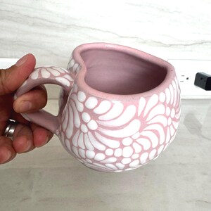 Puede incluir: Una taza de cer&aacute;mica rosa con un dise&ntilde;o floral blanco. La taza tiene un borde en forma de coraz&oacute;n y un asa curva. El interior de la taza es de color rosa liso.