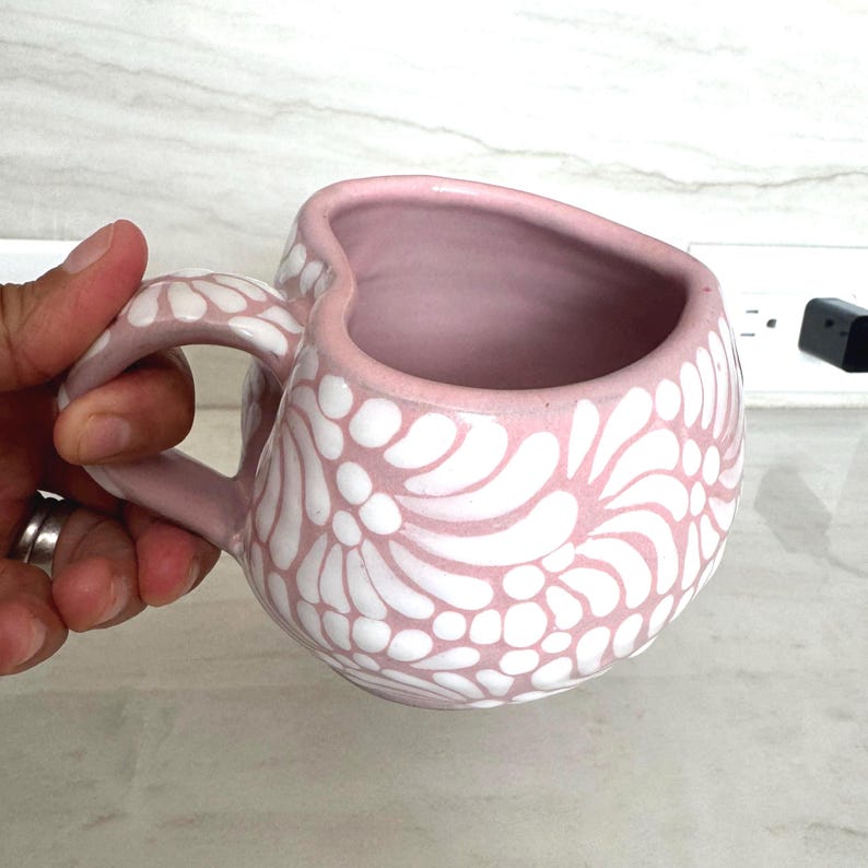 Puede incluir: Una taza de cer&aacute;mica rosa con un dise&ntilde;o floral blanco. La taza tiene un borde en forma de coraz&oacute;n y un asa curva. El interior de la taza es de color rosa liso.