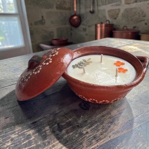 May include: A terracotta clay pot candle with a matching lid, featuring white painted dot patterns. The white wax contains three wicks and is decorated with embedded pressed green leaves and orange flower petals on the surface.