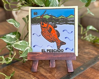 May include: A decorative tile featuring an orange fish illustration with the text "EL PESCADO" on a small wooden easel. The tile has a blue background with water waves and a green and black landscape.