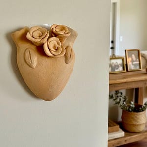 May include: A light brown, heart-shaped wall planter made of unglazed clay. The surface features three sculpted clay roses clustered at the top center, flanked by two vertical, leaf-like indentations on the upper sides of the heart form.