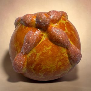 May include: A round, golden-brown bread loaf with a shiny, glazed surface. The top features decorative dough strips arranged in a cross pattern, with small round dough balls at the ends and center, all dusted with a fine layer of sparkling sugar.