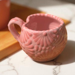 May include: A pink ceramic mug with a heart-shaped rim and a handle. The mug features a textured, embossed floral design. The mug is sitting on a white marble surface. Another mug is partially visible in the background.