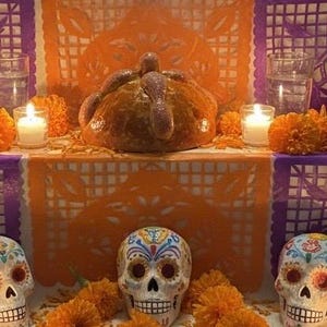 May include: A round, golden-brown pan de muerto bread sits on a tiered altar decorated with orange and purple paper cutouts, bright orange marigold flowers, clear glasses of water, and three white candles, alongside three painted sugar skulls.