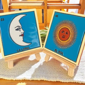 May include: Two decorative ceramic tiles on wooden easels. One tile features a crescent moon with a face, the text "LA LUNA", and the number 23. The other tile depicts a sun with a face, the text "EL SOL", and the number 46. Both tiles have a blue background.