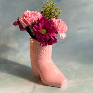 May include: A pink ceramic cowboy boot vase filled with pink carnations, purple chrysanthemums, and green filler flowers. The boot has embossed detailing and a square toe. The background is a light blue and green.