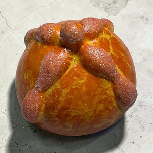 May include: A round, golden-orange decorative object shaped like pan de muerto, featuring a central knob and four radiating bone-like strips across the top. The surface has a glossy, textured finish with a subtle dusting of fine, sparkling glitter.