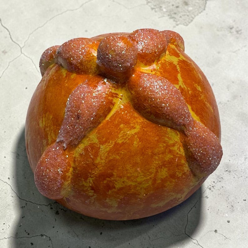 May include: A round, golden-orange decorative object shaped like pan de muerto, featuring a central knob and four radiating bone-like strips across the top. The surface has a glossy, textured finish with a subtle dusting of fine, sparkling glitter.