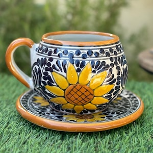 May include: A ceramic teacup and saucer set with a hand-painted sunflower design. The cup and saucer feature a yellow sunflower, black and white floral patterns, and an orange rim and handle. The set is placed on a green surface.