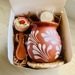 May include: A gift set in a white box, featuring a terracotta mug with white floral designs, a wooden spoon, a round candy with text, and a small candle. The mug sits on a woven coaster, all nestled in brown paper.