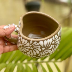 Puede incluir: Una taza de cer&aacute;mica marr&oacute;n con borde y asa en forma de coraz&oacute;n. La taza est&aacute; decorada con motivos florales blancos. El interior de la taza es marr&oacute;n. La taza se sostiene en una mano.