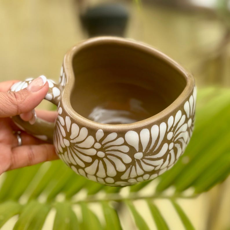 Puede incluir: Una taza de cer&aacute;mica marr&oacute;n con borde y asa en forma de coraz&oacute;n. La taza est&aacute; decorada con motivos florales blancos. El interior de la taza es marr&oacute;n. La taza se sostiene en una mano.