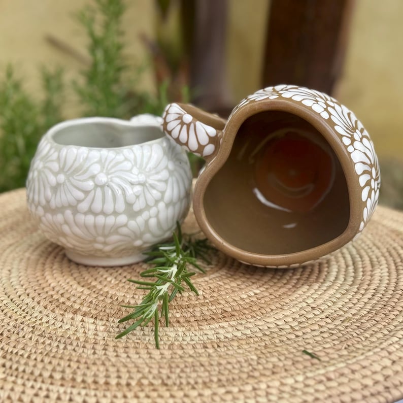 Puede incluir: Dos tazas de cer&aacute;mica con motivos florales. Una taza es de color gris claro con flores blancas, y la otra es marr&oacute;n con flores blancas. Las tazas est&aacute;n sobre una estera tejida con una ramita de romero.