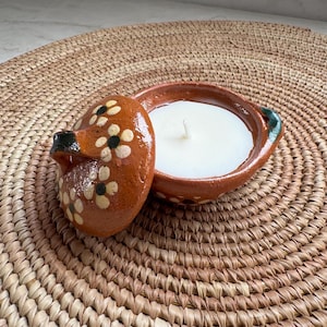 May include: A terracotta clay candle holder with a matching lid, featuring a hand-painted floral pattern with white petals and black centers. The round vessel contains a white wax candle with a centered wick and has a small handle on the side.