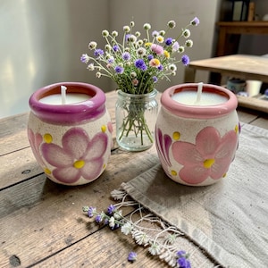 Puede incluir: Dos velas de cerámica en macetas moteadas con diseños florales. Una maceta tiene un borde morado y la otra un borde rosa. Cada vela tiene cera y mecha blancas. Un pequeño jarrón de cristal con flores se encuentra entre las velas.