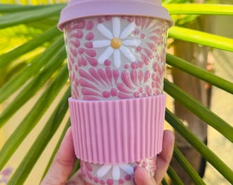 May include: A pink and white floral travel mug with a matching lid and a pink silicone sleeve. The mug features a daisy pattern and is held in front of a green leafy background.