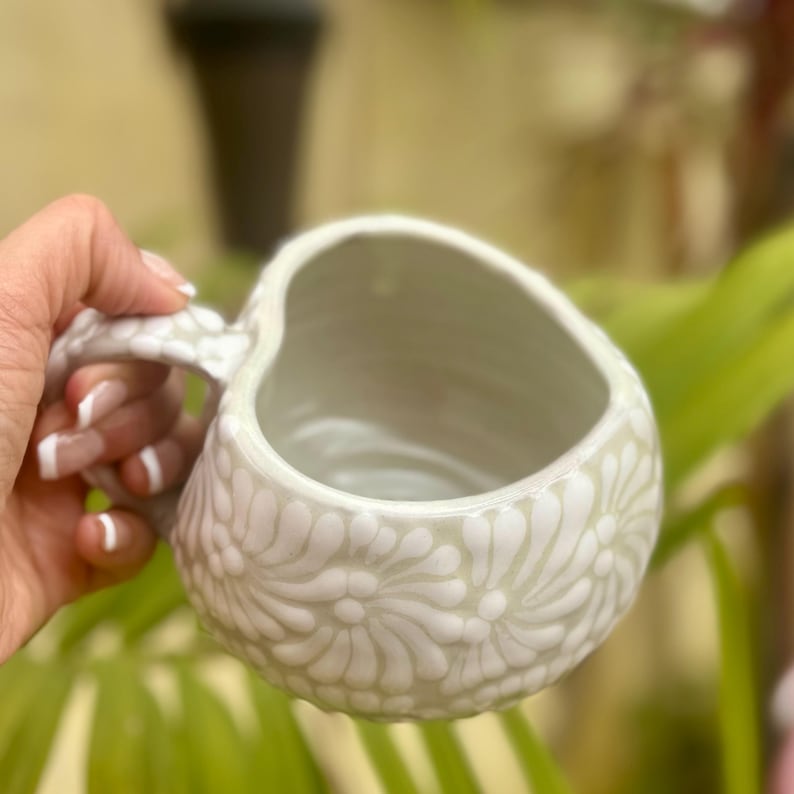 Puede incluir: Una taza de cer&aacute;mica verde claro con un dise&ntilde;o floral. La taza tiene una abertura en forma de coraz&oacute;n y un asa texturizada. El dise&ntilde;o floral es en un tono de verde m&aacute;s claro, creando un contraste sutil. La taza es sostenida por una mano.