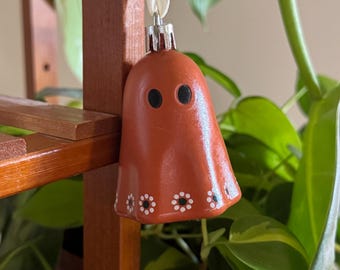 May include: A terracotta-colored ghost-shaped ornament with black eyes and white floral accents. The ornament is suspended by a white ribbon and hangs in front of green foliage. The ornament is a decorative item.