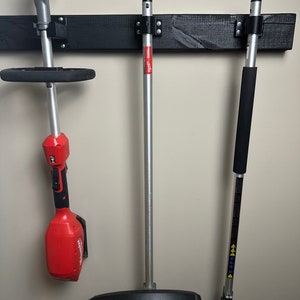 May include: Three black wall-mounted holders with three different types of weed trimmers. The trimmers are all silver and black. The leftmost trimmer is red and black. The middle trimmer has a black and silver handle and a black blade. The rightmost trimmer has a black handle and a black blade.