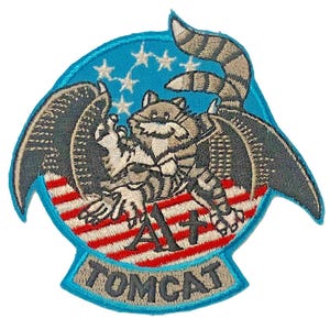 May include: An embroidered patch featuring a cartoon cat with wings and a striped tail, set against a blue background with white stars. Below the cat is a red and white striped pattern with the text A1+ and the word TOMCAT in black lettering.