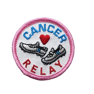 May include: A circular embroidered patch with a pink border and white background. It features two grey running shoes, a red heart, and the blue text CANCER above the shoes and the red text RELAY below the shoes, all stitched in thread.