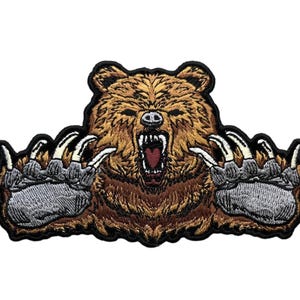 May include: An embroidered patch featuring a brown bear with its mouth open in a roar, displaying sharp white teeth and a dark red tongue. The bear has its two large front paws extended outward, showing detailed grey fur and long white claws.
