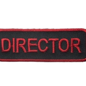 May include: A rectangular embroidered patch with a black background and a red border. The word DIRECTOR is stitched in red capital letters across the center of the patch. The edges of the patch are finished with a red thread border.