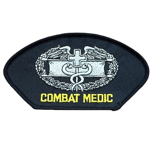 May include: A black, shield-shaped embroidered patch featuring a silver caduceus symbol with a cross and wings, surrounded by a wreath. The text "COMBAT MEDIC" is stitched in yellow capital letters at the bottom of the patch, which has a black border.