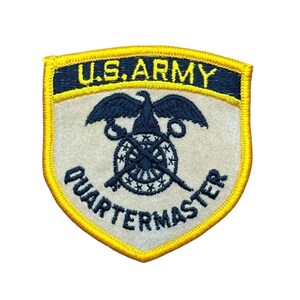 May include: A shield-shaped embroidered patch with a yellow border. The top dark blue section features the yellow text U.S. ARMY. The light beige center displays a dark blue eagle emblem above a crossed sword and key, with the word QUARTERMASTER below.