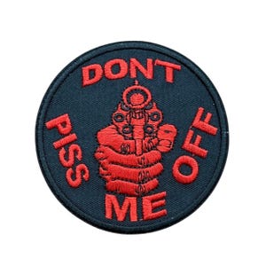 May include: A circular embroidered patch with a black background and red stitching. The design features a hand holding a revolver pointed forward, surrounded by the red text DON'T PISS ME OFF arranged in a circular pattern around the central graphic.