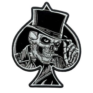May include: An embroidered patch in the shape of a spade playing card suit. The design features a skull wearing a black top hat and a dark jacket, with one skeletal hand touching the brim of the hat. The embroidery uses black, white, and gray thread.